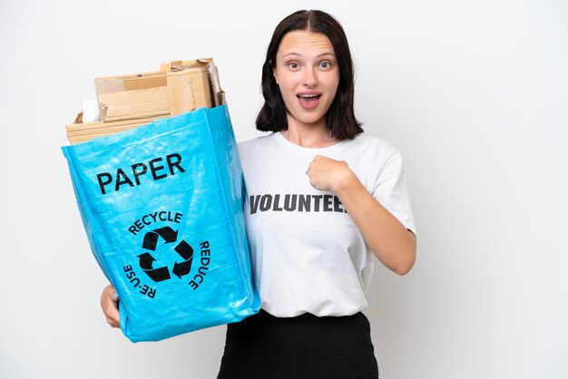 Jeune femme caucasienne tenant un sac de recyclage plein de papier à recycler isolé sur fond blanc avec une expression faciale surprise