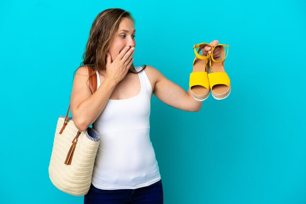 Jeune femme caucasienne tenant des sandales d'été isolées sur fond bleu avec une expression faciale surprise et choquée