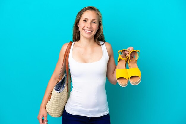 Jeune femme caucasienne tenant des sandales d'été isolées sur fond bleu souriant beaucoup