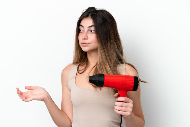 Jeune femme caucasienne tenant un sèche-cheveux isolé sur fond blanc avec une expression faciale surprise