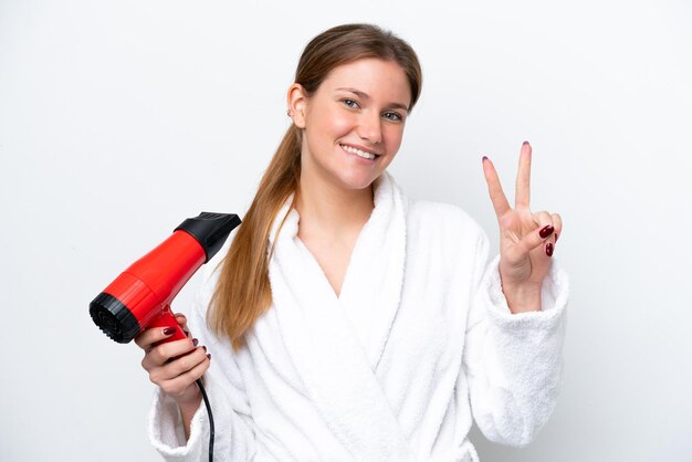 Jeune femme caucasienne tenant un sèche-cheveux isolé sur fond blanc souriant et montrant le signe de la victoire