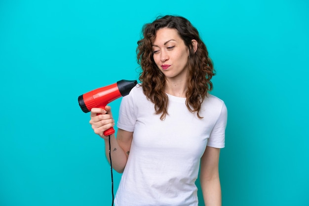 Jeune femme caucasienne tenant un sèche-cheveux isolé sur fond bleu avec une expression triste