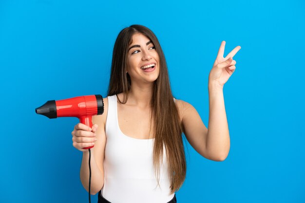 Jeune femme caucasienne tenant un sèche-cheveux isolé sur fond bleu souriant et montrant le signe de la victoire