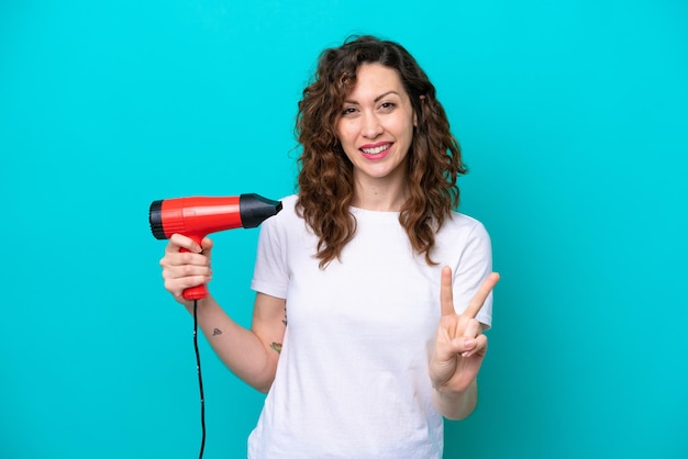 Jeune femme caucasienne tenant un sèche-cheveux isolé sur fond bleu souriant et montrant le signe de la victoire