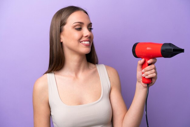 Jeune femme caucasienne tenant un sèche-cheveux isolé sur fond violet avec une expression heureuse