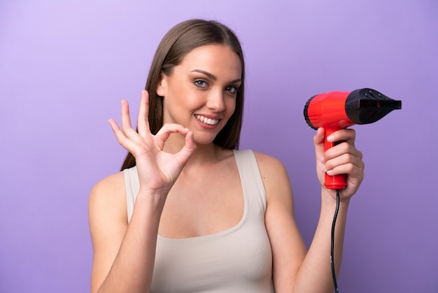 Jeune femme caucasienne tenant un sèche-cheveux isolé sur fond violet montrant un signe ok avec les doigts