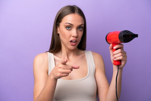 Jeune femme caucasienne tenant un sèche-cheveux isolé sur fond violet surpris et pointant vers l'avant