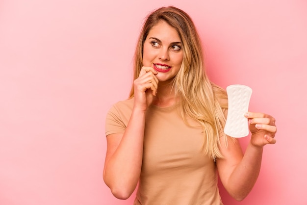 Jeune femme caucasienne tenant une serviette hygiénique isolée sur fond rose détendu en pensant à quelque chose en regardant un espace de copie