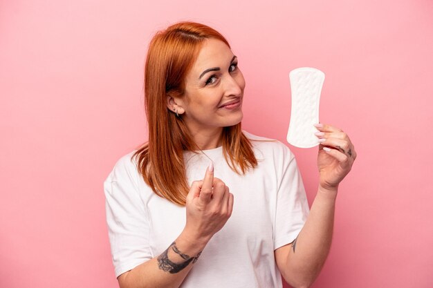 Photo jeune femme caucasienne tenant une serviette hygiénique isolée sur fond rose pointant du doigt vers vous comme si vous invitiez à vous rapprocher.