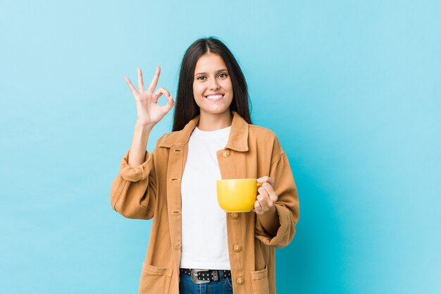 Jeune femme caucasienne tenant une tasse de café gaie et confiante, montrant le geste ok.