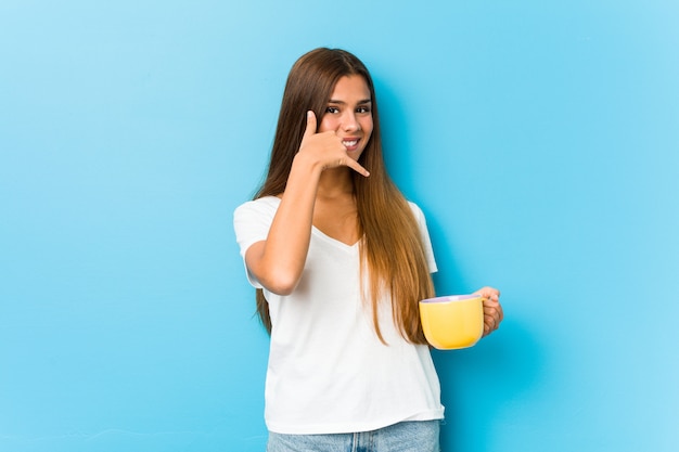 Jeune femme caucasienne tenant une tasse de café montrant un geste d'appel de téléphone mobile avec les doigts.