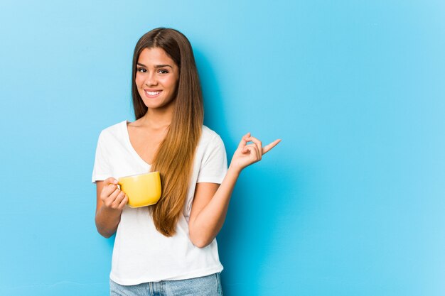 Jeune femme caucasienne tenant une tasse de café en souriant et en pointant de côté, montrant quelque chose à l'espace vide.