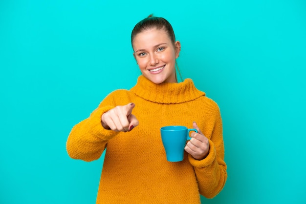 Jeune femme caucasienne tenant une tasse isolée sur fond bleu pointe le doigt vers vous avec une expression confiante