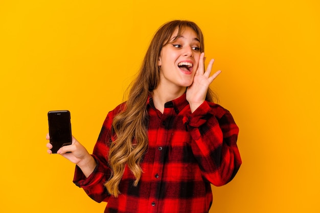 Jeune femme caucasienne tenant le téléphone isolé sur fond jaune criant et tenant la paume près de la bouche ouverte.