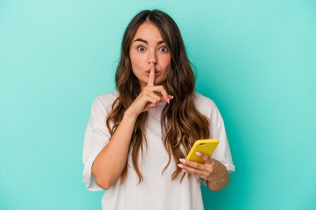 Jeune femme caucasienne tenant un téléphone portable isolé sur fond bleu gardant un secret ou demandant le silence.