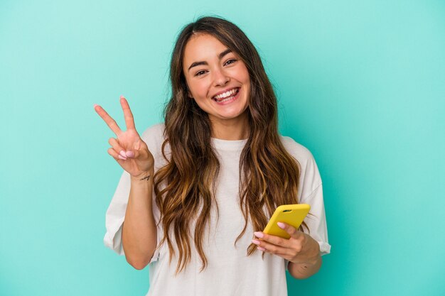 Jeune femme caucasienne tenant un téléphone portable isolé sur fond bleu joyeux et insouciant montrant un symbole de paix avec les doigts.