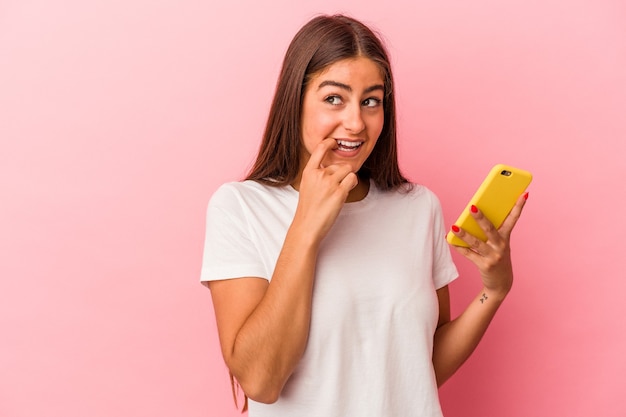 Jeune femme caucasienne tenant un téléphone portable isolé sur fond rose détendue en pensant à quelque chose en regardant un espace de copie.