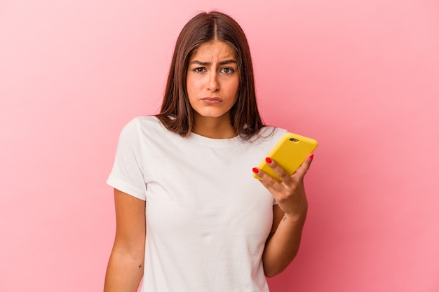 Jeune femme caucasienne tenant un téléphone portable isolé sur fond rose hausse les épaules et ouvre les yeux confus.