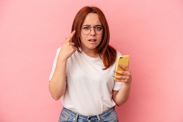 Jeune femme caucasienne tenant un téléphone portable isolé sur fond rose montrant un geste de déception avec l'index.