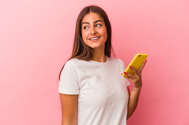 Jeune femme caucasienne tenant un téléphone portable isolé sur fond rose regarde de côté souriante, gaie et agréable.