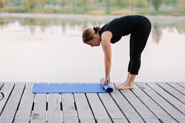 Jeune femme caucasienne tord le tapis de yoga à l'extérieur près de la rivière sur une jetée en bois le matin