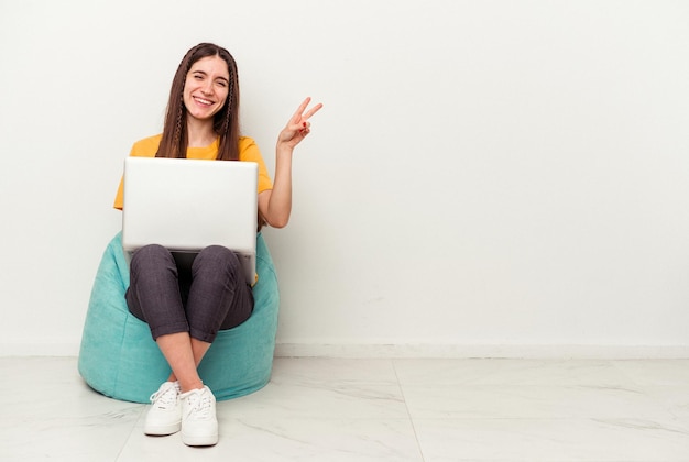 Jeune femme caucasienne travaillant avec un ordinateur portable assis sur une bouffée isolé sur fond blanc joyeux et insouciant montrant un symbole de paix avec les doigts