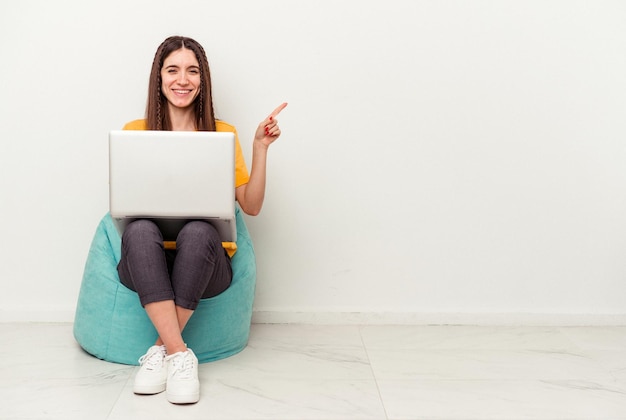 Jeune femme caucasienne travaillant avec un ordinateur portable assis sur une bouffée isolé sur fond blanc souriant et pointant de côté montrant quelque chose à l'espace vide