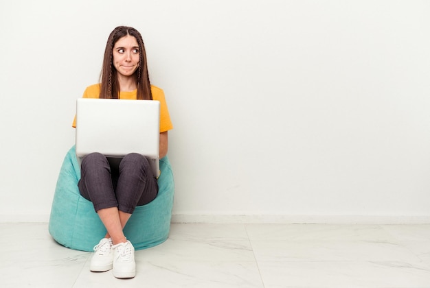 Jeune femme caucasienne travaillant avec un ordinateur portable assis sur une bouffée isolée sur fond blanc confus se sent douteux et incertain