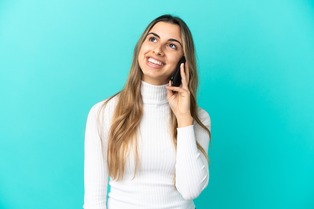 Jeune femme caucasienne utilisant un téléphone portable isolé sur fond bleu en pensant à une idée tout en levant les yeux