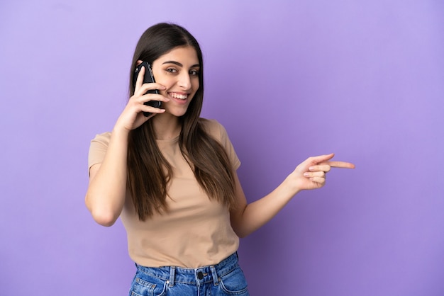 Jeune femme caucasienne utilisant un téléphone portable isolé sur fond violet, pointant le doigt sur le côté