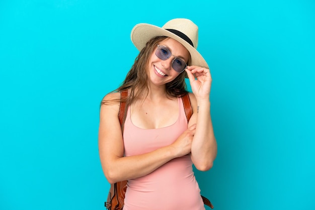 Jeune femme caucasienne en vacances d'été isolée sur fond bleu avec des lunettes et heureuse