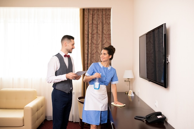 Jeune femme de chambre avec détergent et plumeau et porteur avec pavé tactile interagissant dans l'une des chambres d'hôtel au travail