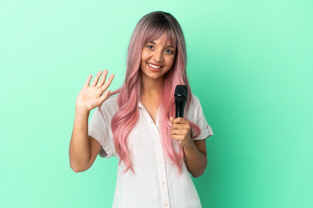 Jeune femme chanteuse métisse aux cheveux roses isolée sur fond vert saluant avec la main avec une expression heureuse