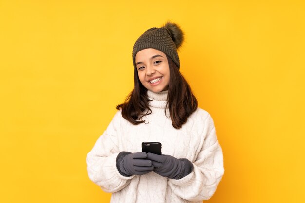 Jeune femme avec chapeau d'hiver fond jaune isolé envoyant un message avec le mobile