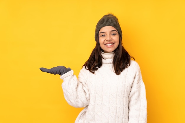 Jeune femme avec chapeau d'hiver sur fond jaune isolé tenant imaginaire sur la paume pour insérer une annonce