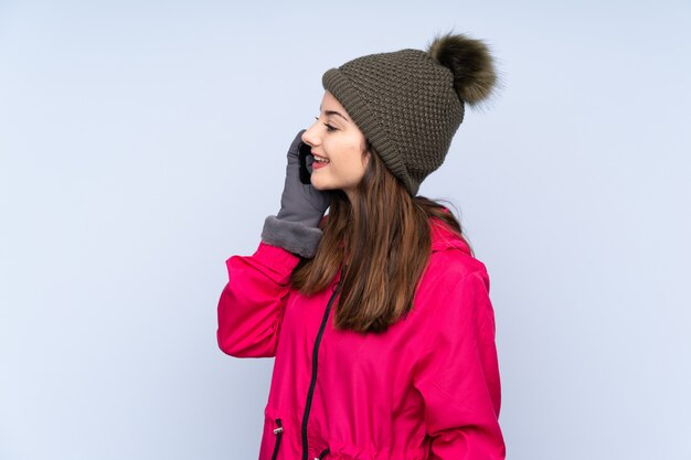 Jeune femme avec un chapeau d'hiver isolé sur le mur bleu en gardant une conversation avec le téléphone mobile avec quelqu'un