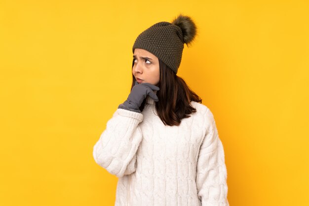 Jeune femme avec un chapeau d'hiver sur un mur jaune isolé ayant des doutes et avec une expression de visage confuse