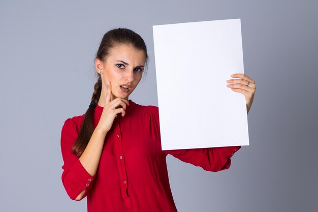 Jeune femme charmante tenant une feuille de papier blanche et pensant sur fond gris en studio