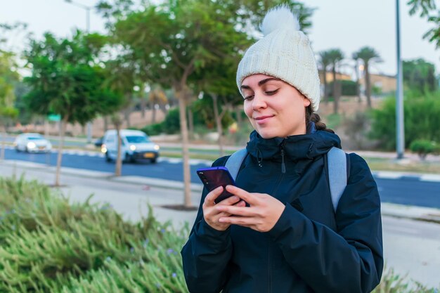 Une jeune femme chaude avec une veste et un chapeau de laine utilise son téléphone portable alors qu'elle se promène dans un parc de la ville