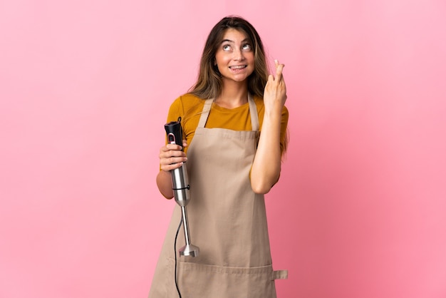 Jeune femme chef à l'aide d'un mixeur à main isolé sur un mur rose avec des doigts qui se croisent et souhaitant le meilleur