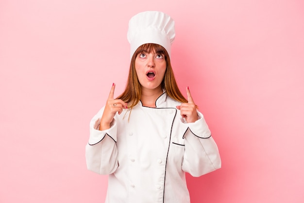 Jeune femme chef caucasienne isolée sur fond rose pointant vers le haut avec la bouche ouverte.