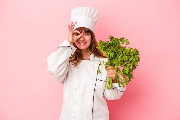 Jeune femme chef caucasienne tenant du persil isolé sur fond rose excité en gardant le geste ok sur les yeux.