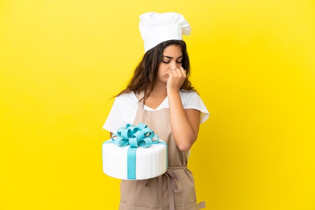 Jeune femme de chef pâtissier caucasien avec un gros gâteau isolé sur fond jaune ayant des doutes