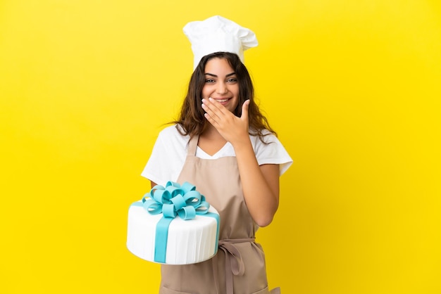 Jeune femme de chef pâtissier caucasien avec un gros gâteau isolé sur fond jaune heureux et souriant couvrant la bouche avec la main