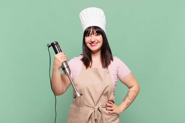 Jeune femme chef souriante joyeusement avec une main sur la hanche et une attitude confiante, positive, fière et amicale