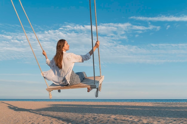 Une jeune femme en chemise blanche sur une corde se balançait sur la mer sur une plage de sable bleu avec des nuages en arrière-plan