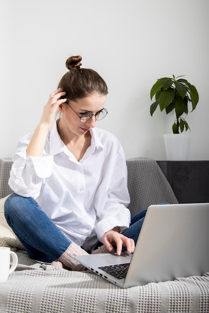 Jeune femme en chemise blanche avec un ordinateur portable travaillant assis sur le canapé