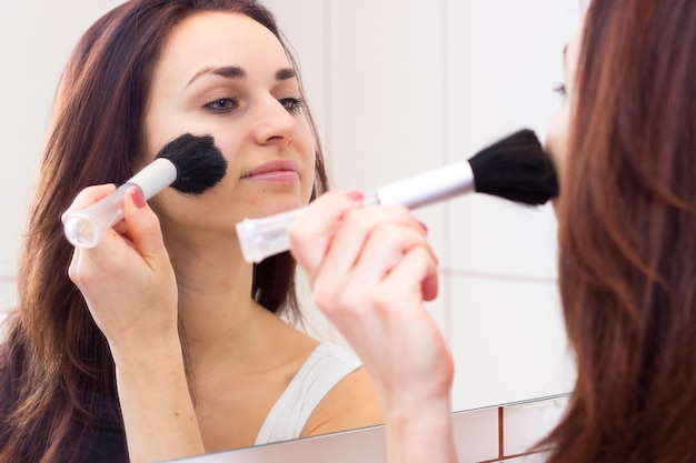 Jeune femme en chemise blanche poudrant avec un pinceau devant le miroir de sa salle de bain bordeaux