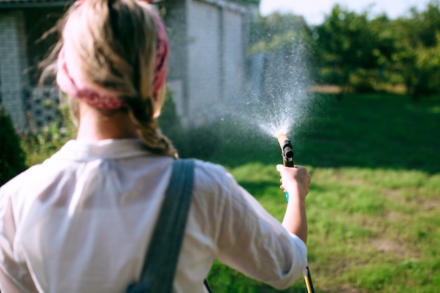 Une jeune femme en chemise blanche et salopette en jean arrosant la pelouse. concept de jardinage et aménagement paysager