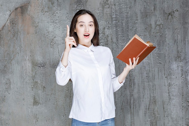 Jeune femme en chemise blanche tenant un livre pour sa classe et pointant. Photo de haute qualité